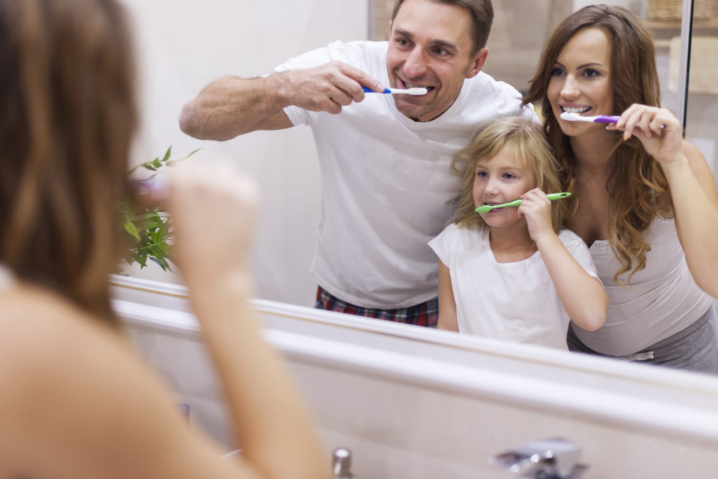 mother, father and child happily brushing their teeth together in the mirror of a well lit bathroom